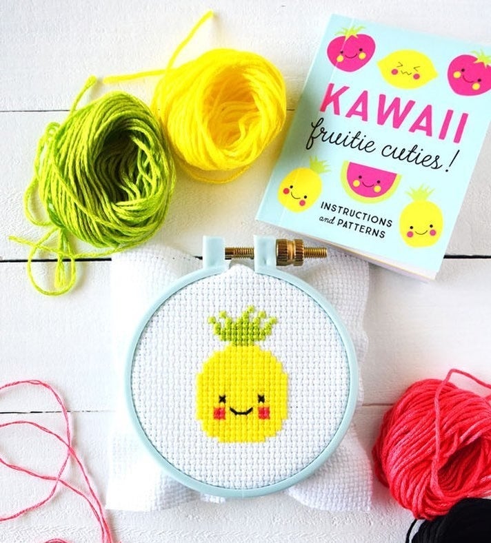 A completed smiling pineapple cross stitch design in a hoop next to thread and the instruction manual