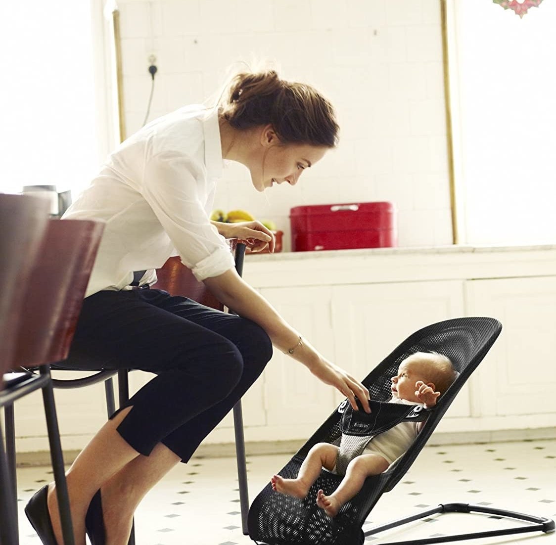 A person sitting on a chair and looking at their baby The baby is sitting upright in a mesh lounge seat on the floor