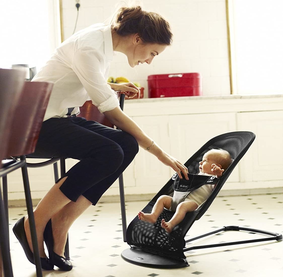 A person sitting on a chair and looking at their baby The baby is sitting upright in a mesh lounge seat on the floor