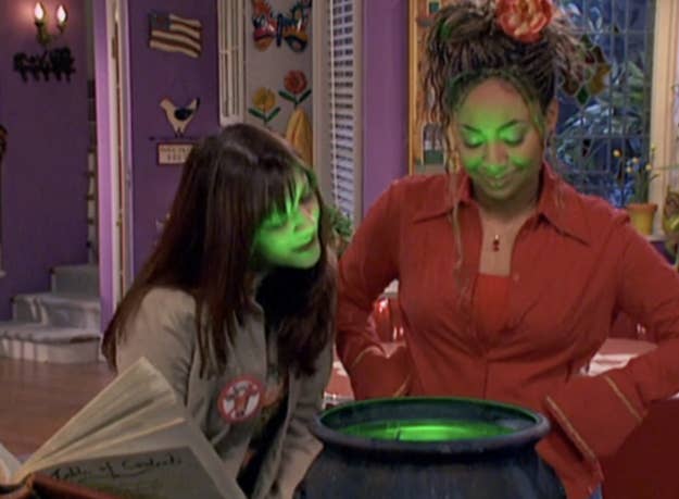 Raven and Chelsea learning over a cauldron next to a spell book. A bright green glow illuminates their faces.