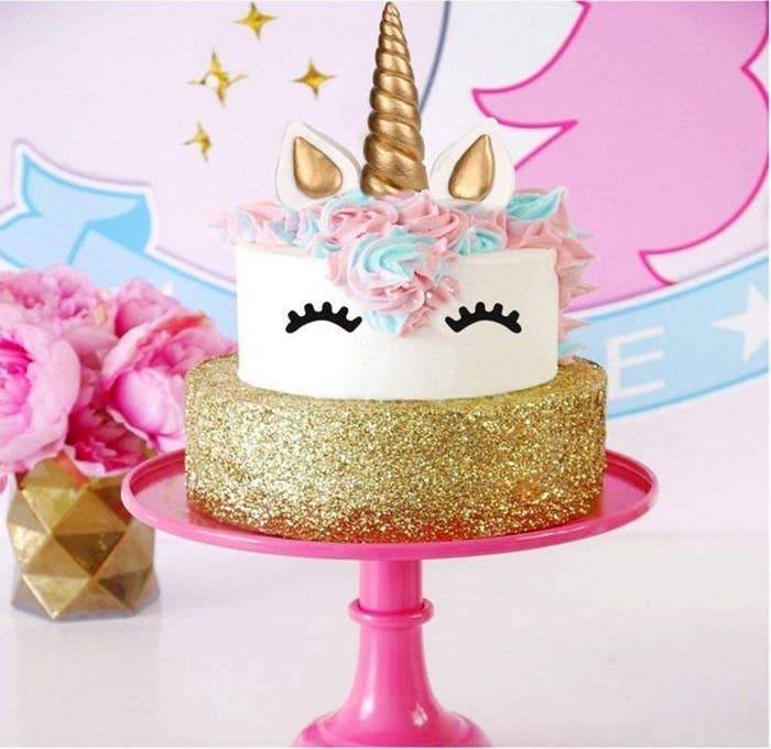 A two-tier cake on a glossy stand with the unicorn cake topper set applied to the top tier