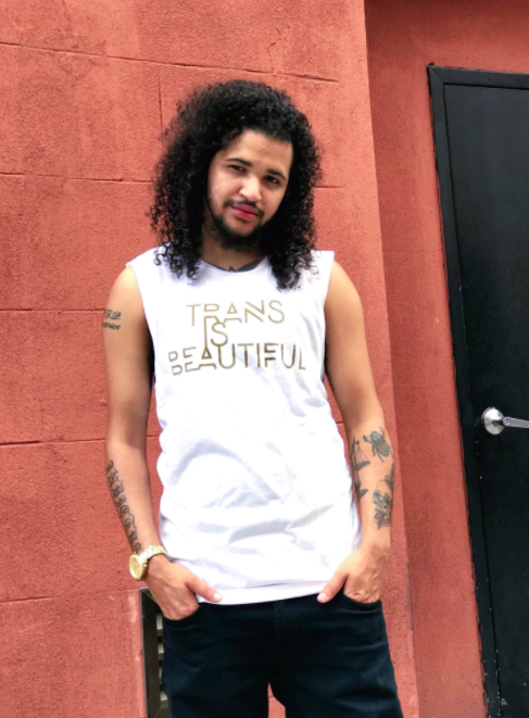 Model wears white tank top with gold letters that say &quot;Trans Is Beautiful&quot;