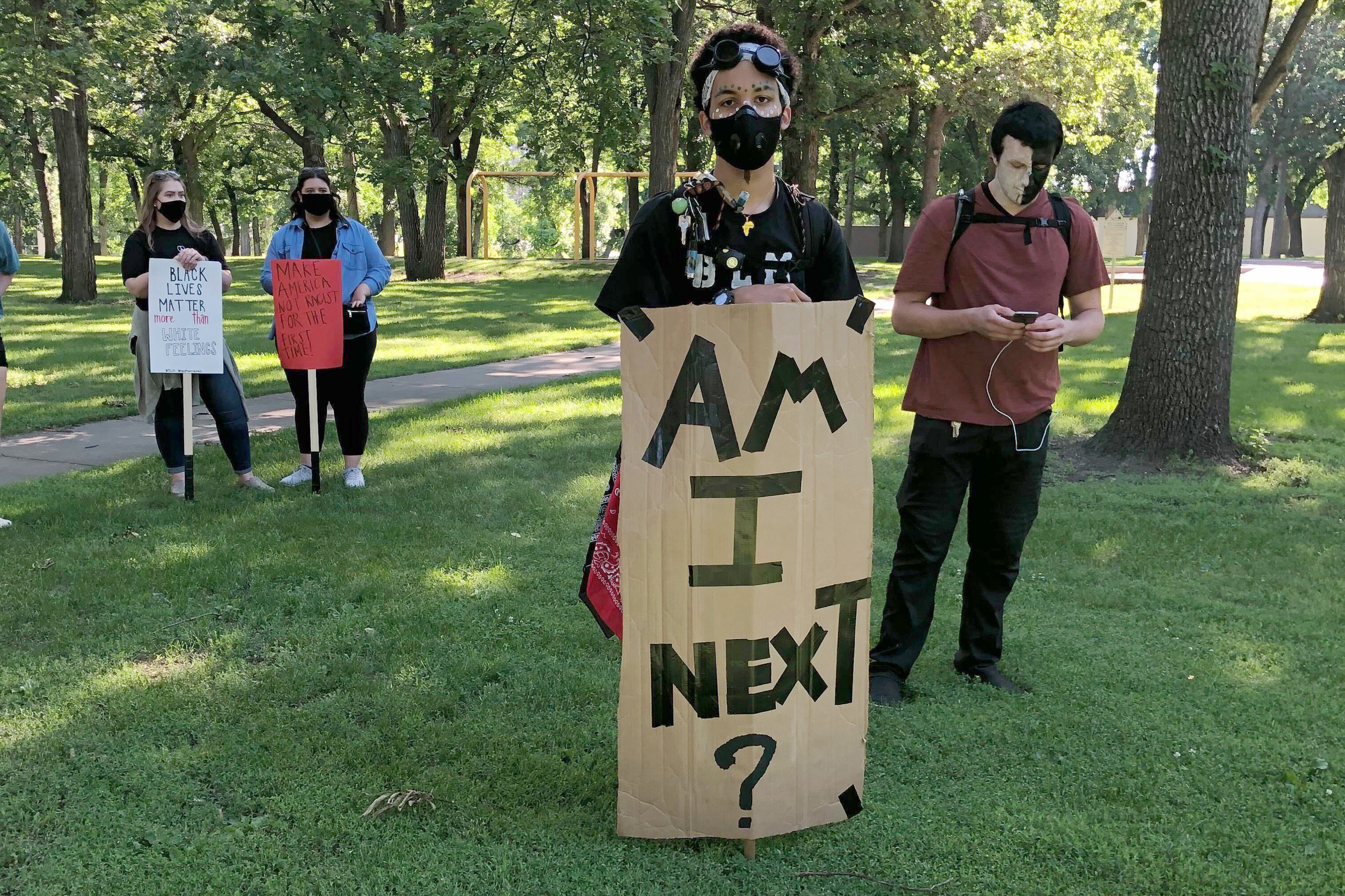 Four people stand in a park. A young person holds a sign that reads, &quot;Am I Next?&quot;