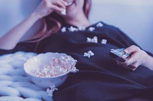 A woman sitting on her bed in her pyjamas, eating popcorn and watching a movie.