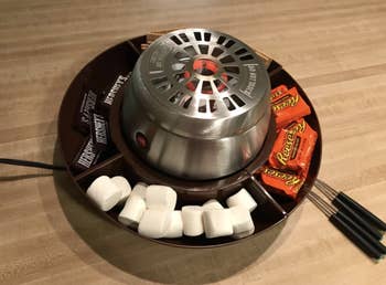 A s'mores maker with marshmallows, Reese's, and graham crackers around it 