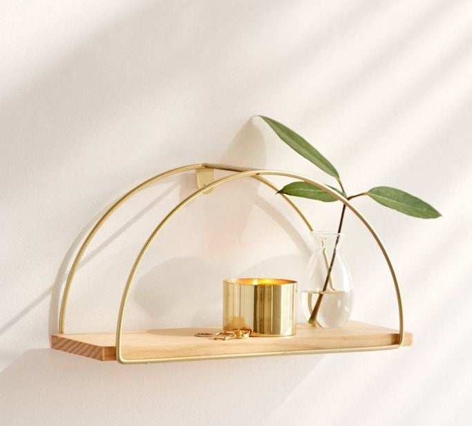 A lifestyle shot of the shelf, made with one piece of wood and two arched, metal decorative pieces, holding a candle, a few rings, and a small bottled plant