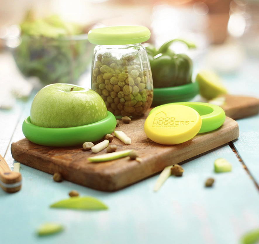 Green reusable food huggers on half an apple and jar of capers