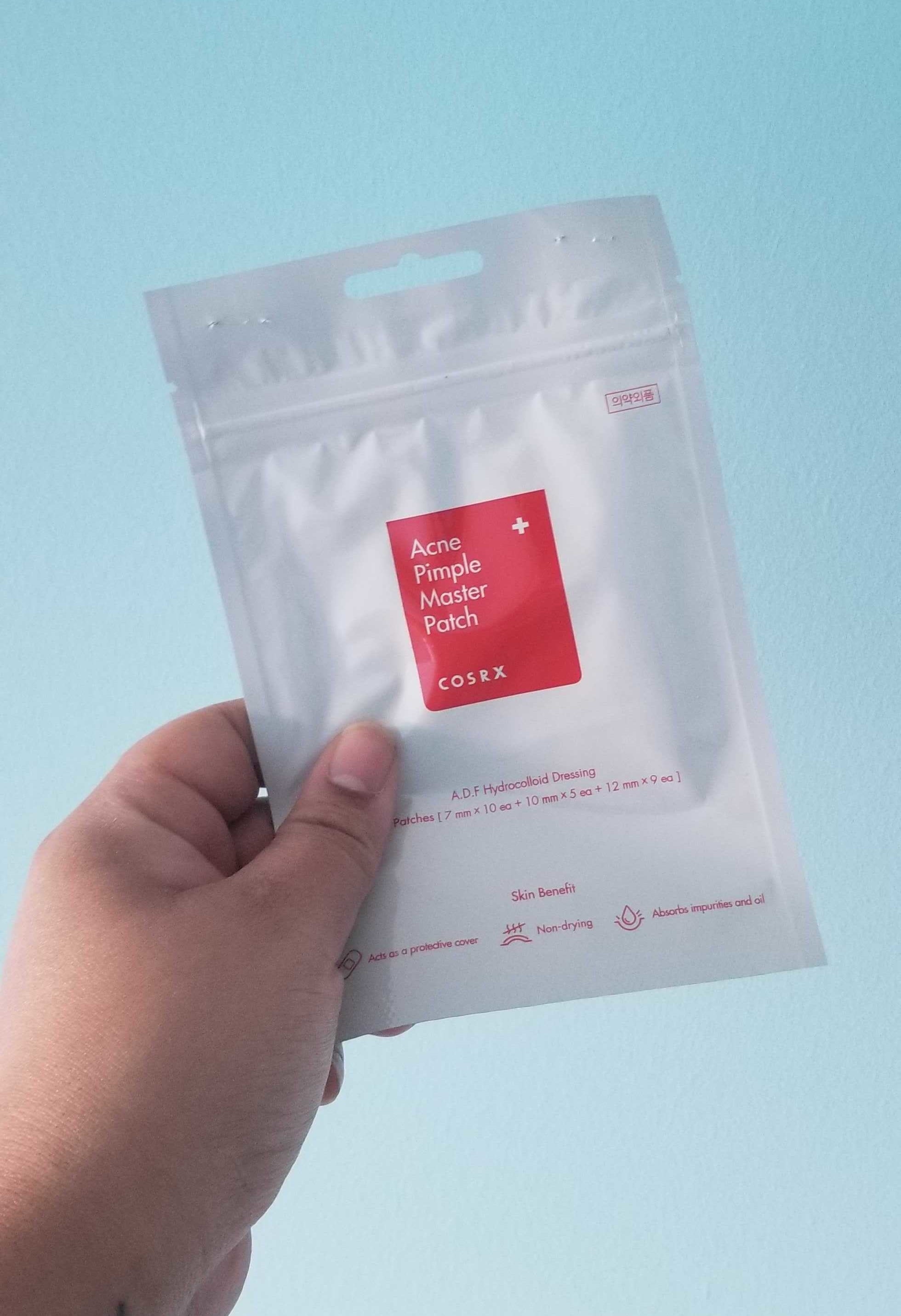A hand holds a small package of acne patches against a wall