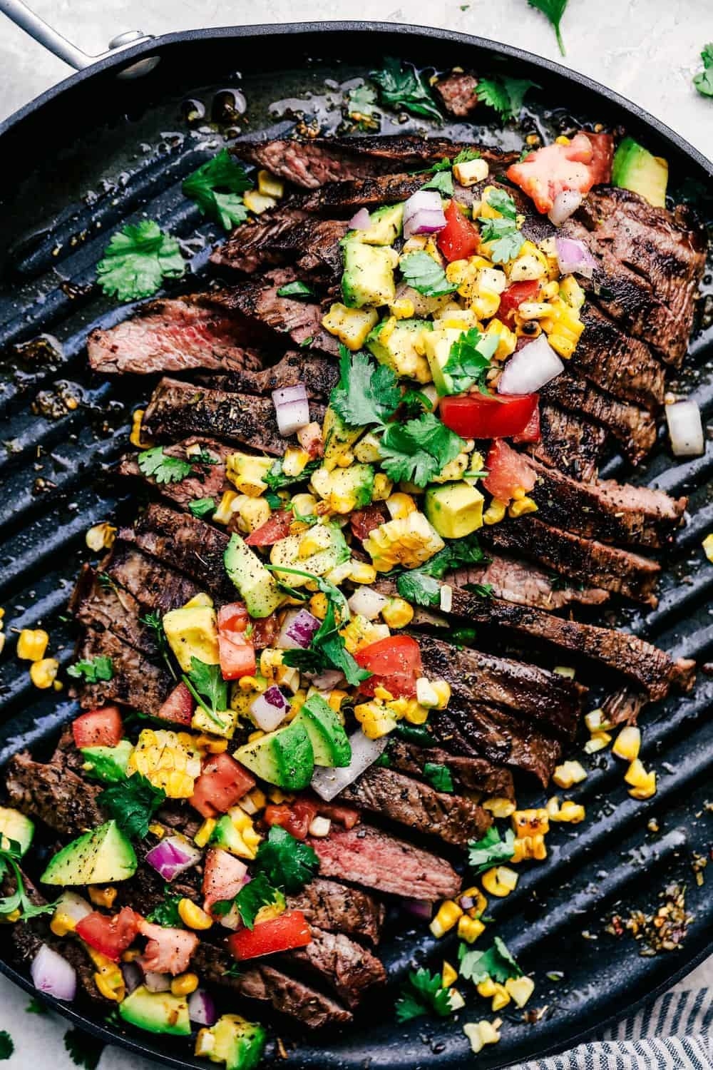 Sliced flank steak on a grill topped with avocado, red onion, tomato, corn, and cilantro slaw.