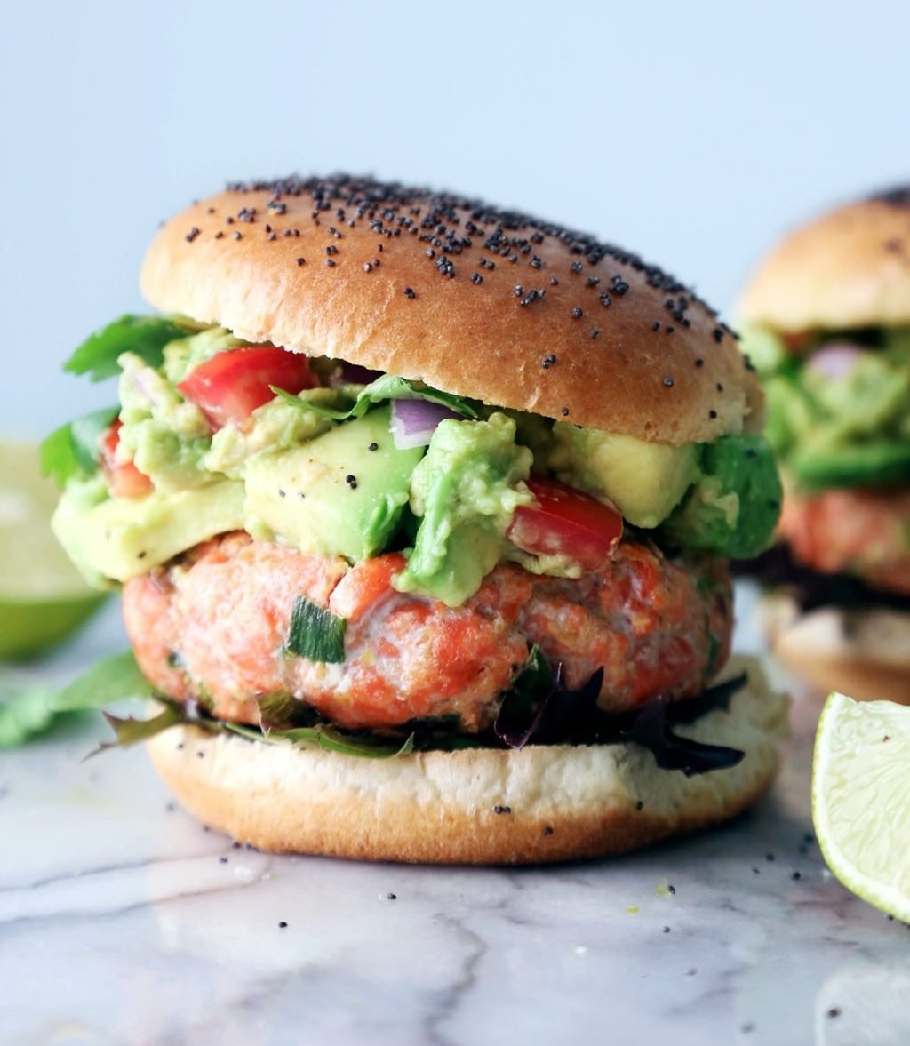 A hamburger bun filled with a thick salmon patty topped with chunky guacamole and diced tomatoes.