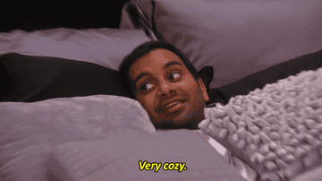 Aziz Ansari as Tom Haverford in Parks And Rec, covered in pillows, saying &quot;very cozy&quot;