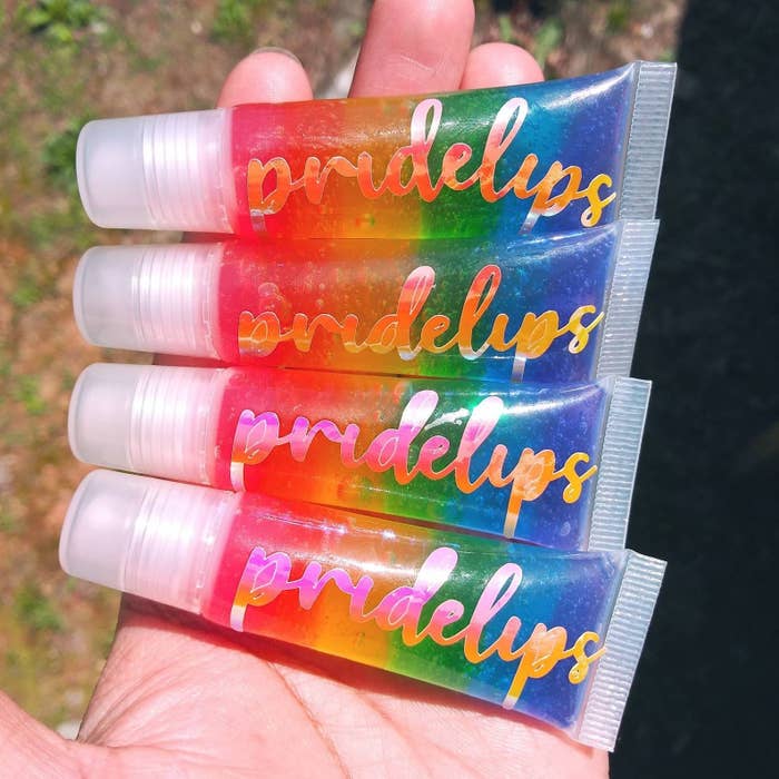 Four tubes of lip gloss with rainbow gloss inside and "pridelips" printed on the front in a model's hands