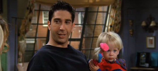 Cena de Ross Geller na série. Ele veste uma blusa azul na casa de Mônica e segura seu filho, que por sua vez, está brincando com uma Barbie.