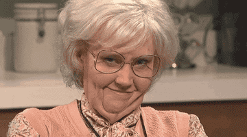 Kate McKinnon grins and raises her eyebrows dressed up as an old lady
