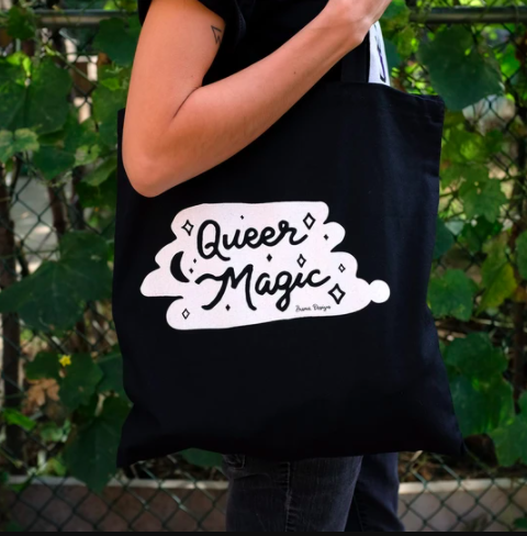 A black tote bag with an illustration on the front in white that says Queer Magic
