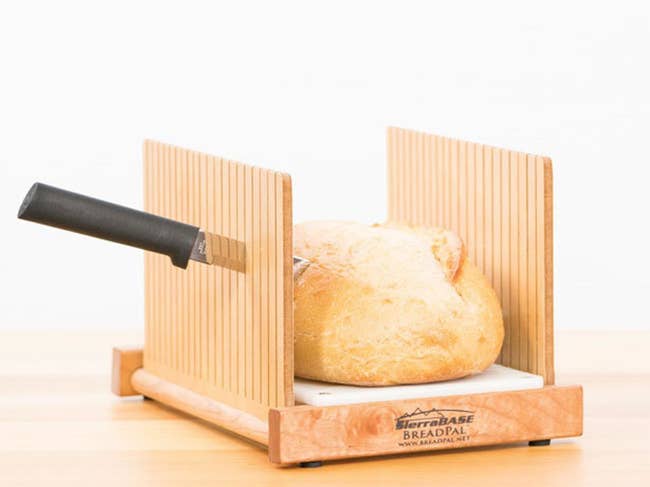 The wooden bread slicer, with a knife cutting between the slats and through the loaf