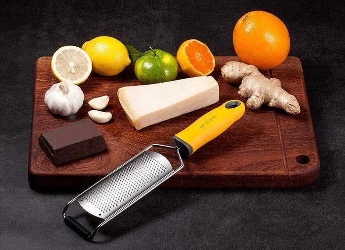 A grater is on a cutting board with cheese, chocolate, and fruit