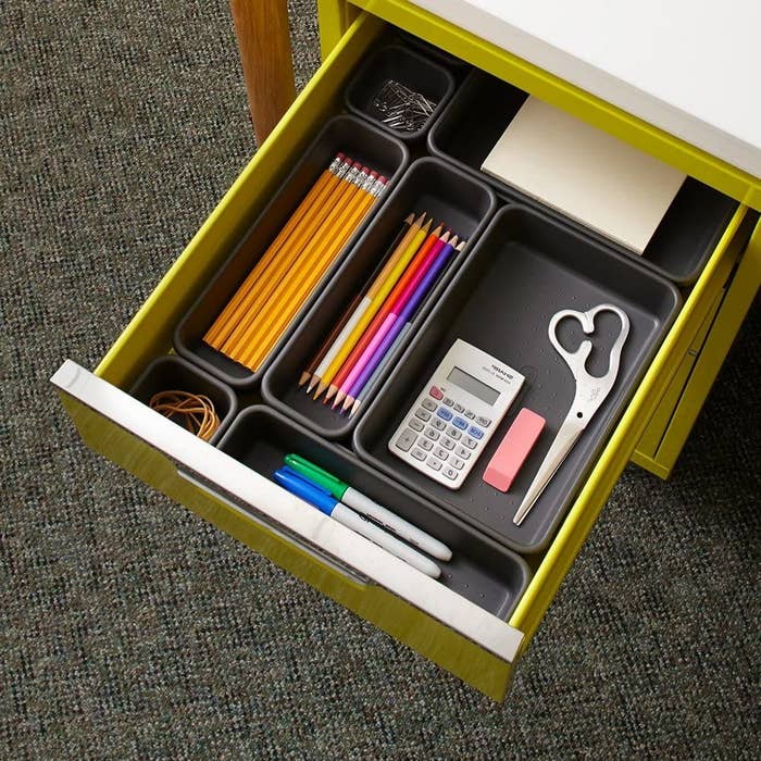 A open desk drawer with containers of different sizes Office supplies like pencils, scissors, and elastic bands are organized in each bin