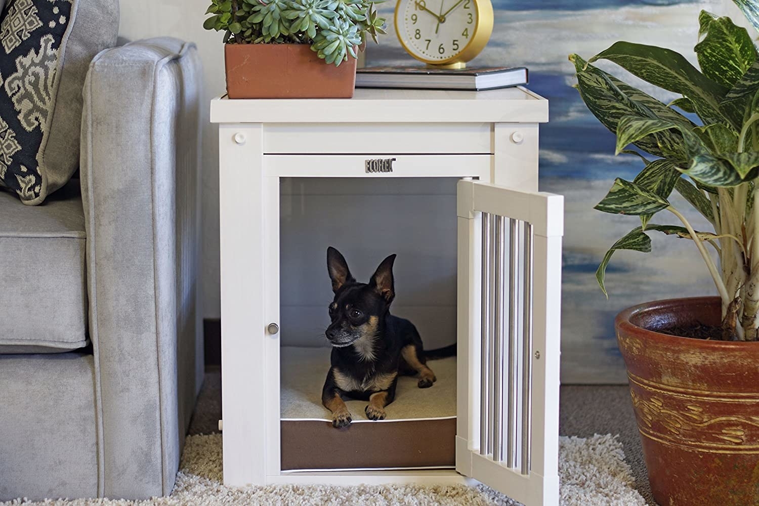 A lifestyle shot of a chiwawa laying inside of the side table crate with the door open. The table is beside a sofa and has a plant, clock, and book as decor on top.