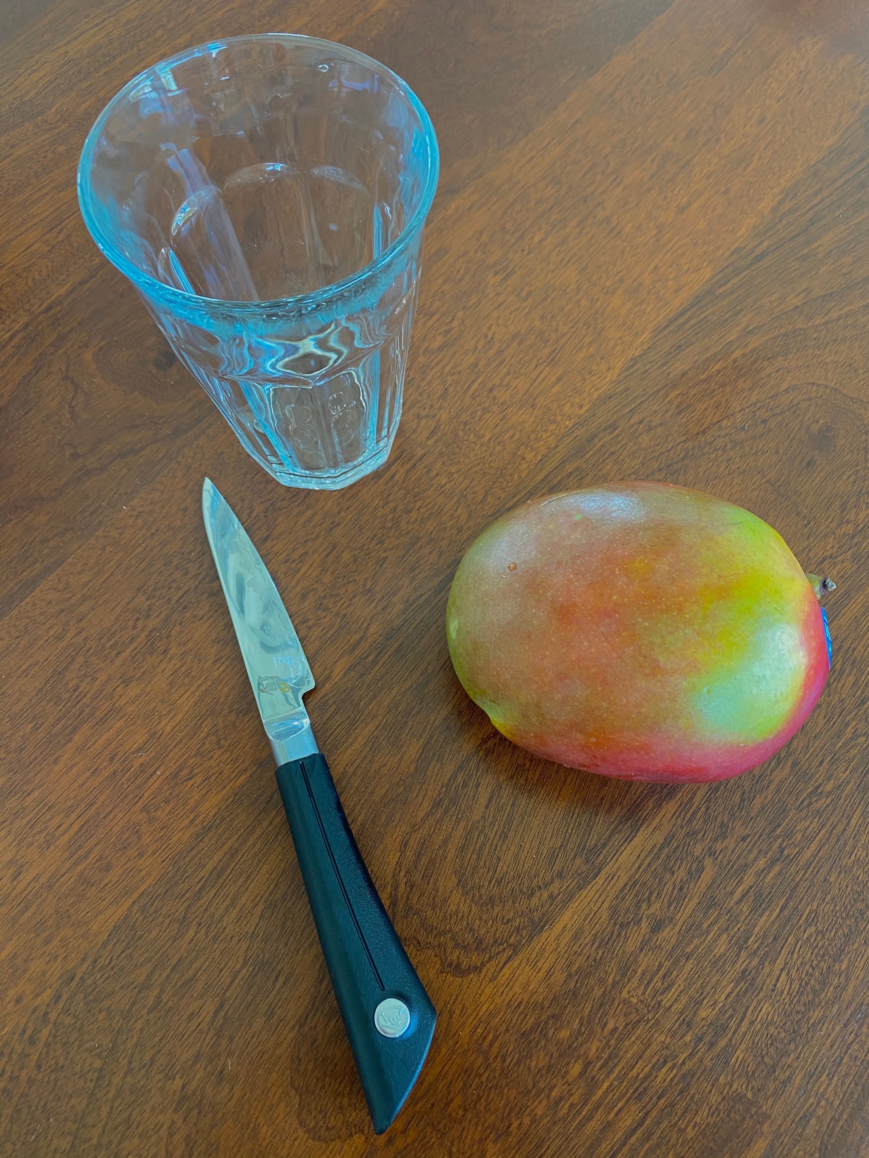 A knife, a whole mango, and a drinking glass on a wooden countertop.