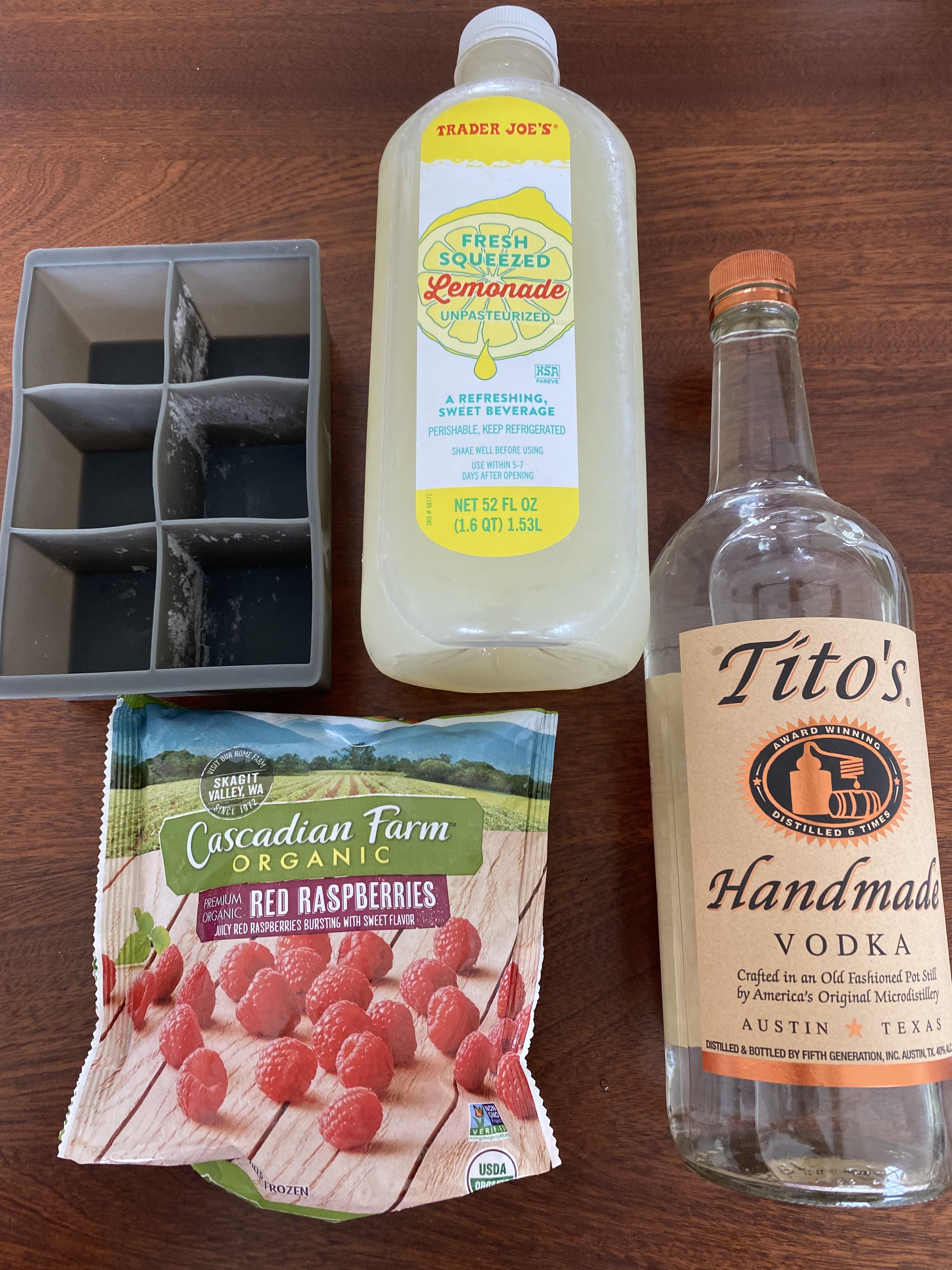 A bottle of Tito&#x27;s vodka, frozen raspberries, a bottle of lemonade, and ice cube molds on a countertop.
