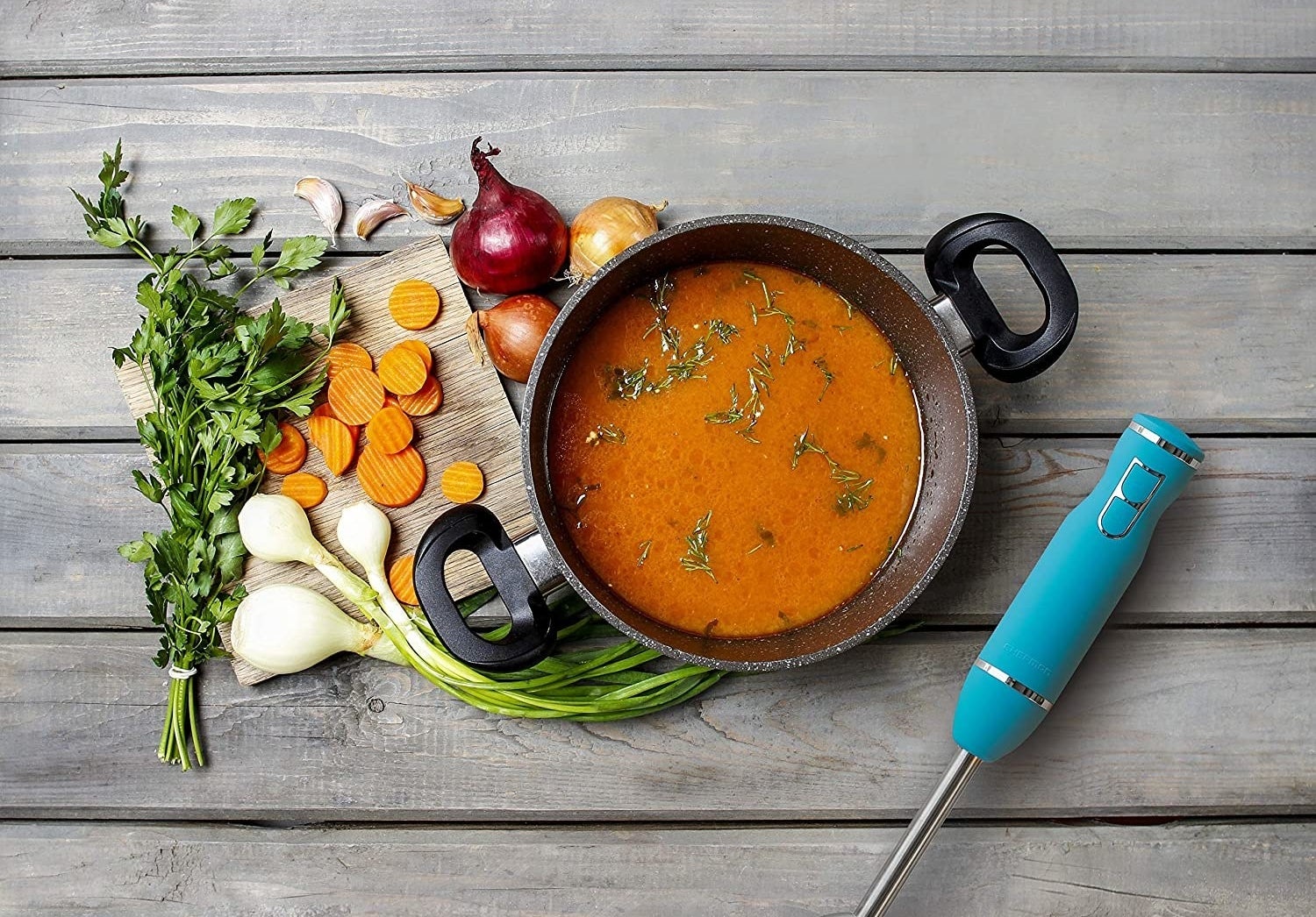 An immersion blender beside a pot of soup and with veggies on a cutting board