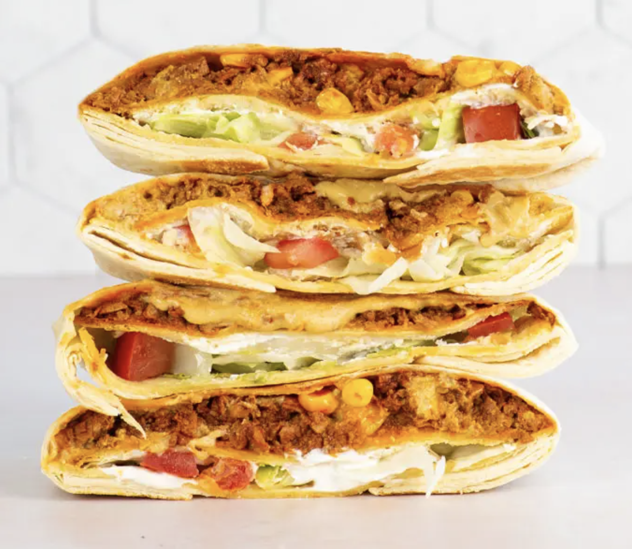 Four stacked burritos are halved to show the filling of seasoned beef, lettuce, tomato, corn, and cheese. The background appears to be a light-colored countertop