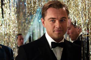 Leonardo DiCaprio as Jay Gatsby wears a tuxedo, looking slightly to the left of the frame. He stands in front of gold streamers.