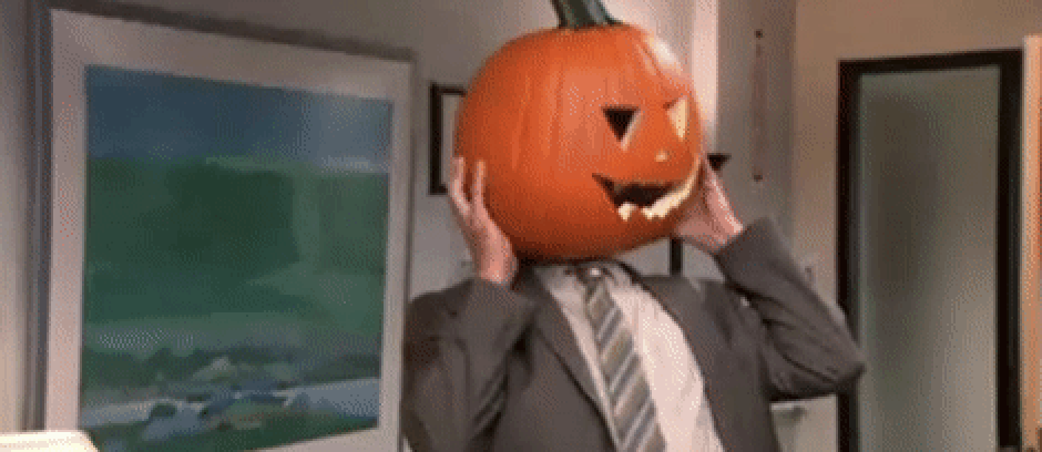 Dwight Schrute from The Office is wearing a gray suit with a white shirt and gray striped tie. On his head is a jack-o-lantern and he is struggling to get it off.