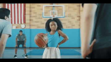 A little girl wearing a blue top and white skirt is facing away from a basketball net. She throws it behind her without looking, and the ball goes right into the net.