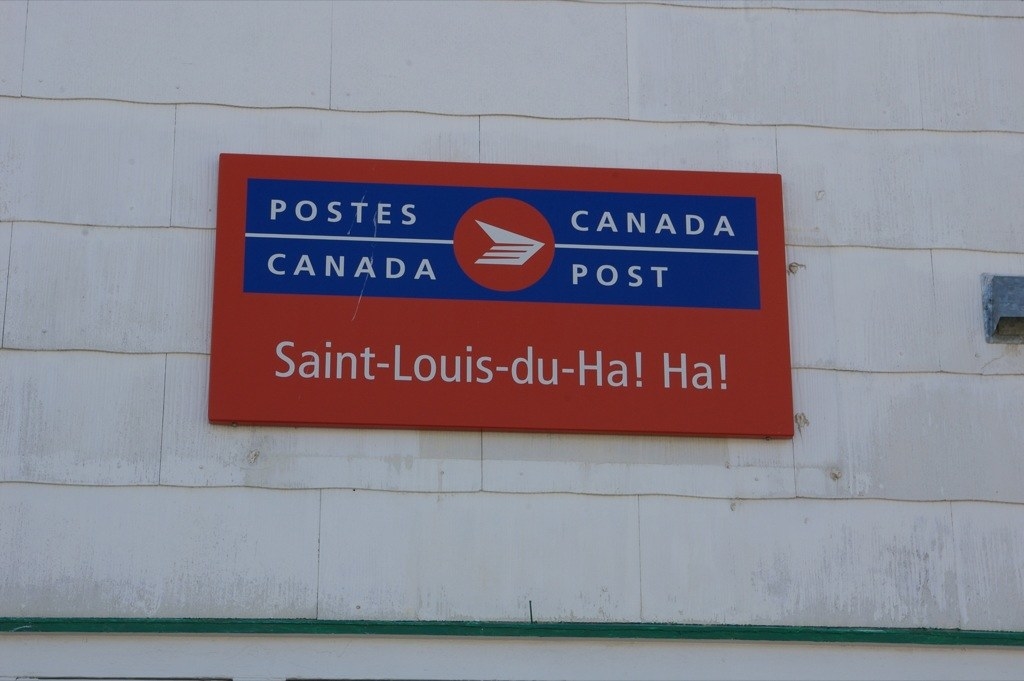 A post office sign that is red and blue reads, &quot;postes Canada/Canada post&quot; on top. Below it, the words Saint-Louis-du-Ha! Ha!