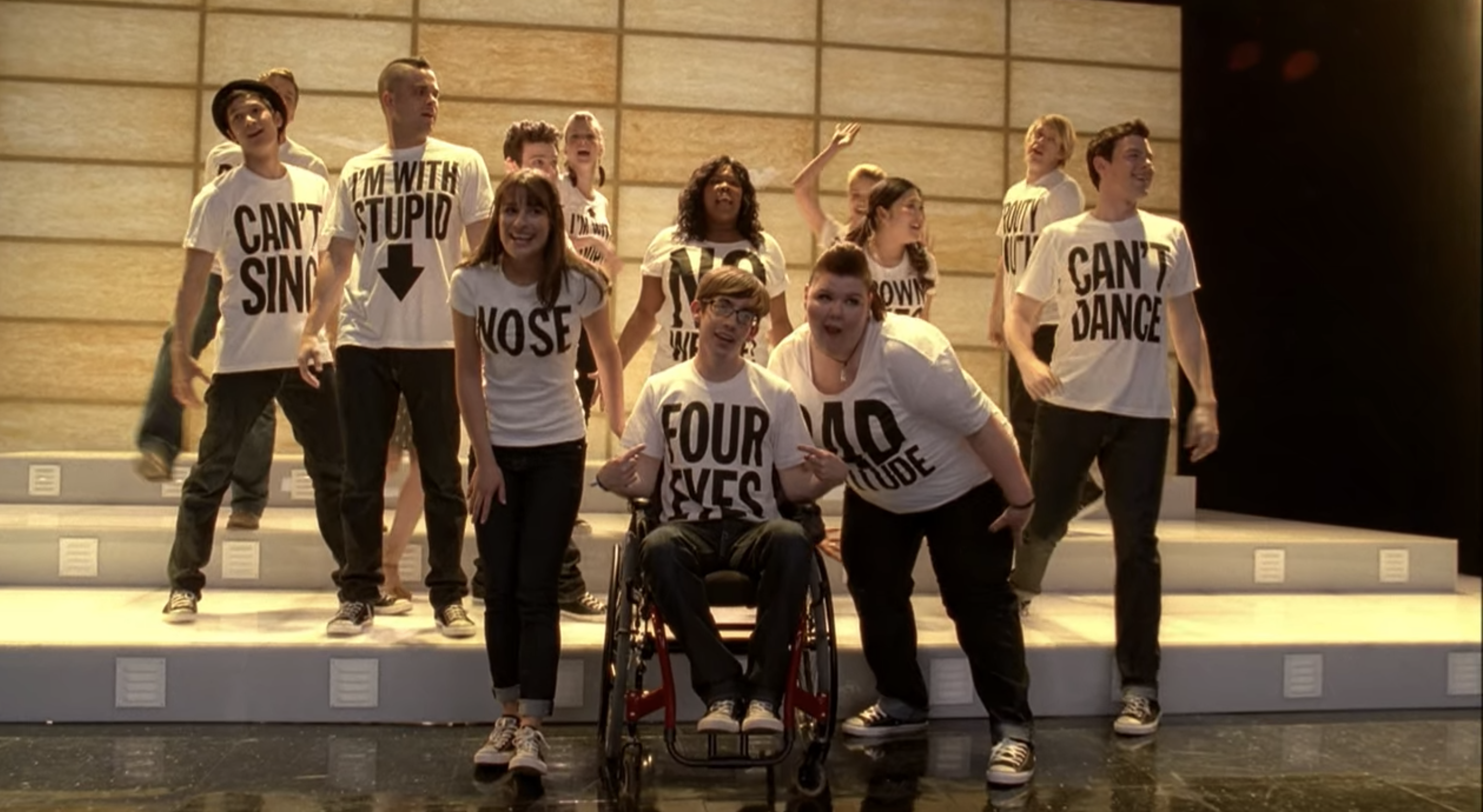 The New Directions sing on stage while wearing white T-shirts that state the individual traits they are proud of.