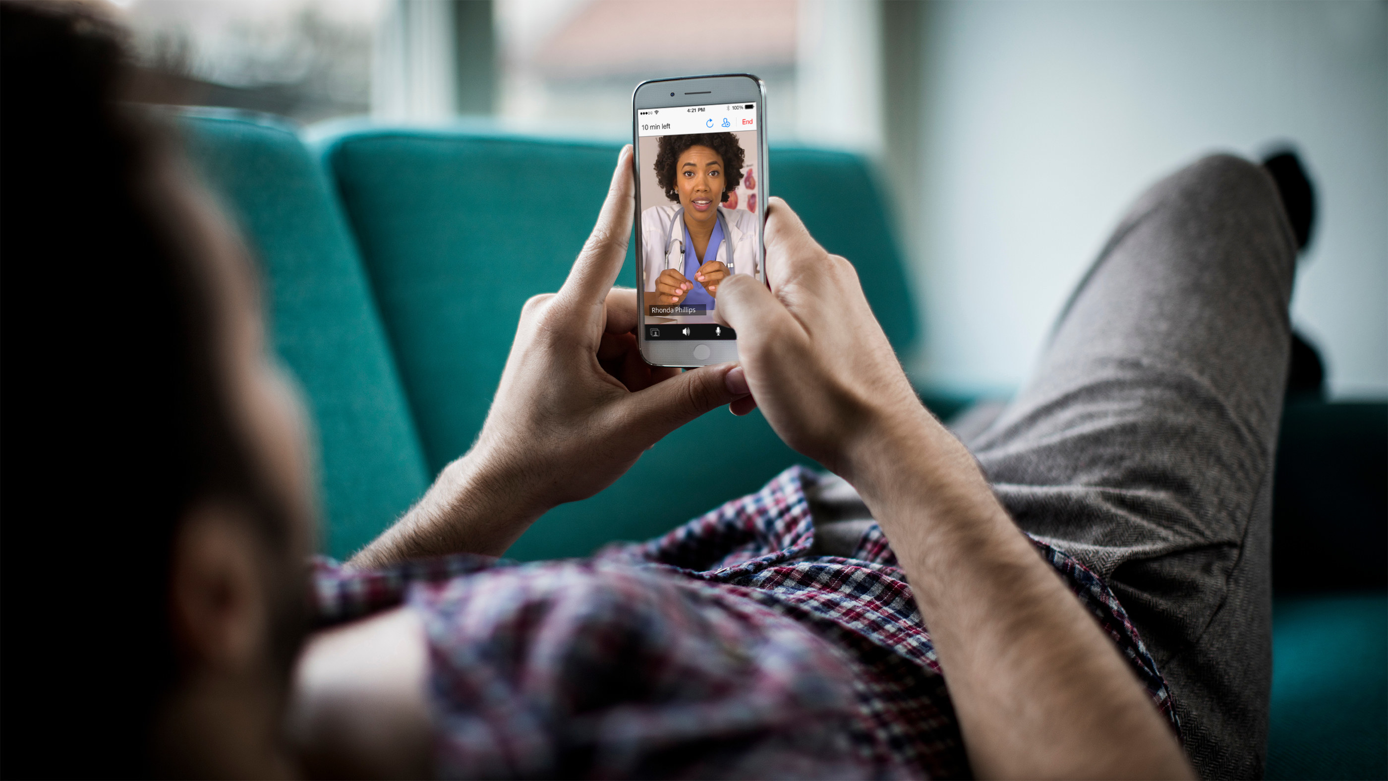 A woman lies on her couch and holds up her smartphone as she video conferences her therapist