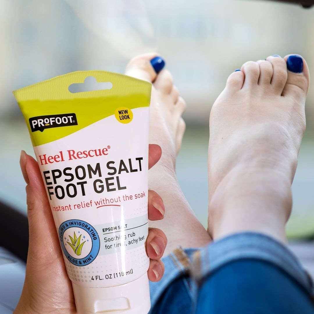A person holding a tube of epsom salt foot gel in front of their feet