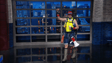 A person wearing head-to-toe camping gear and giving a thumbs-up on The Late Show With Stephen Colbert 