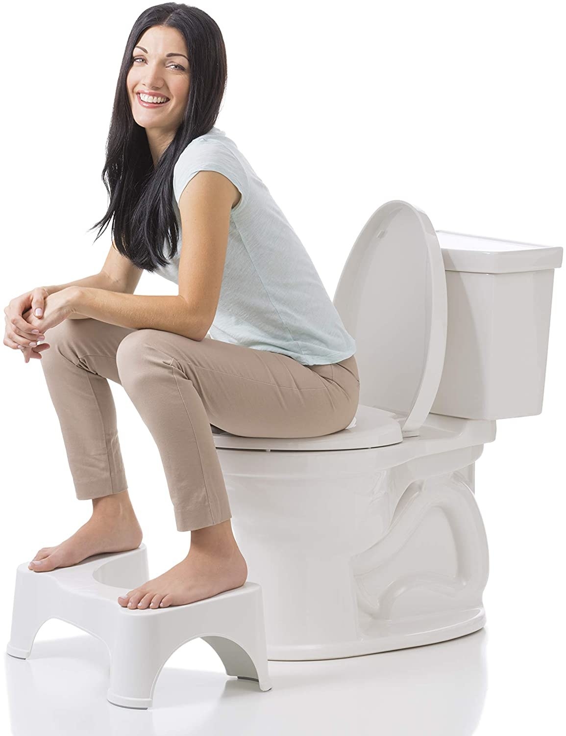 A model sitting on the toilet using the Squatty Potty, which is white and comes up to about half the height of the toilet. The model has their feet on either side of the stool