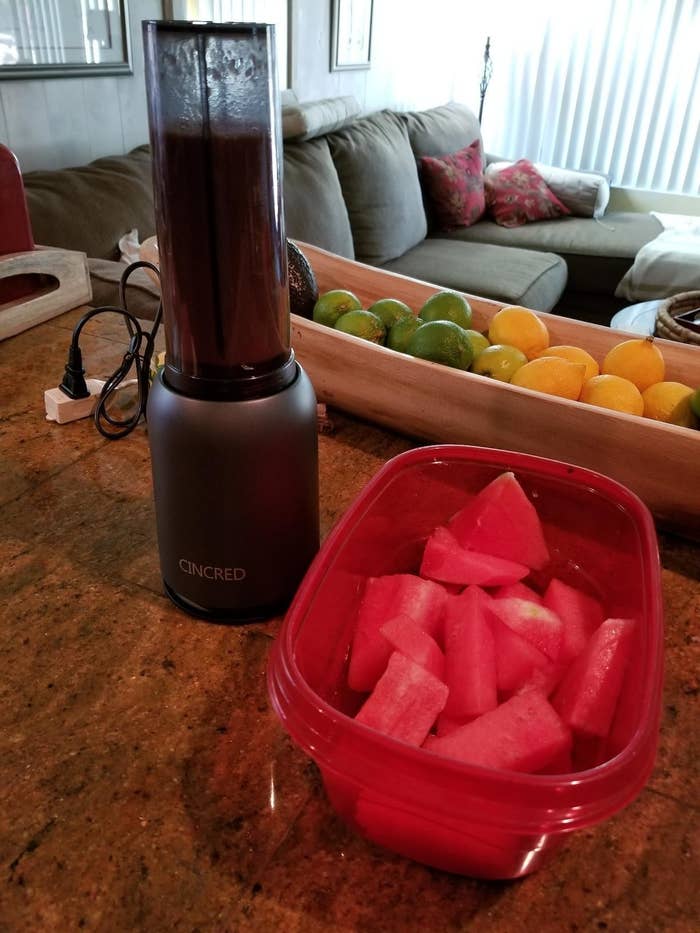 A reviewer&#x27;s photo of a blender with a metallic gray base and a clear plastic cup filled with blended fruit