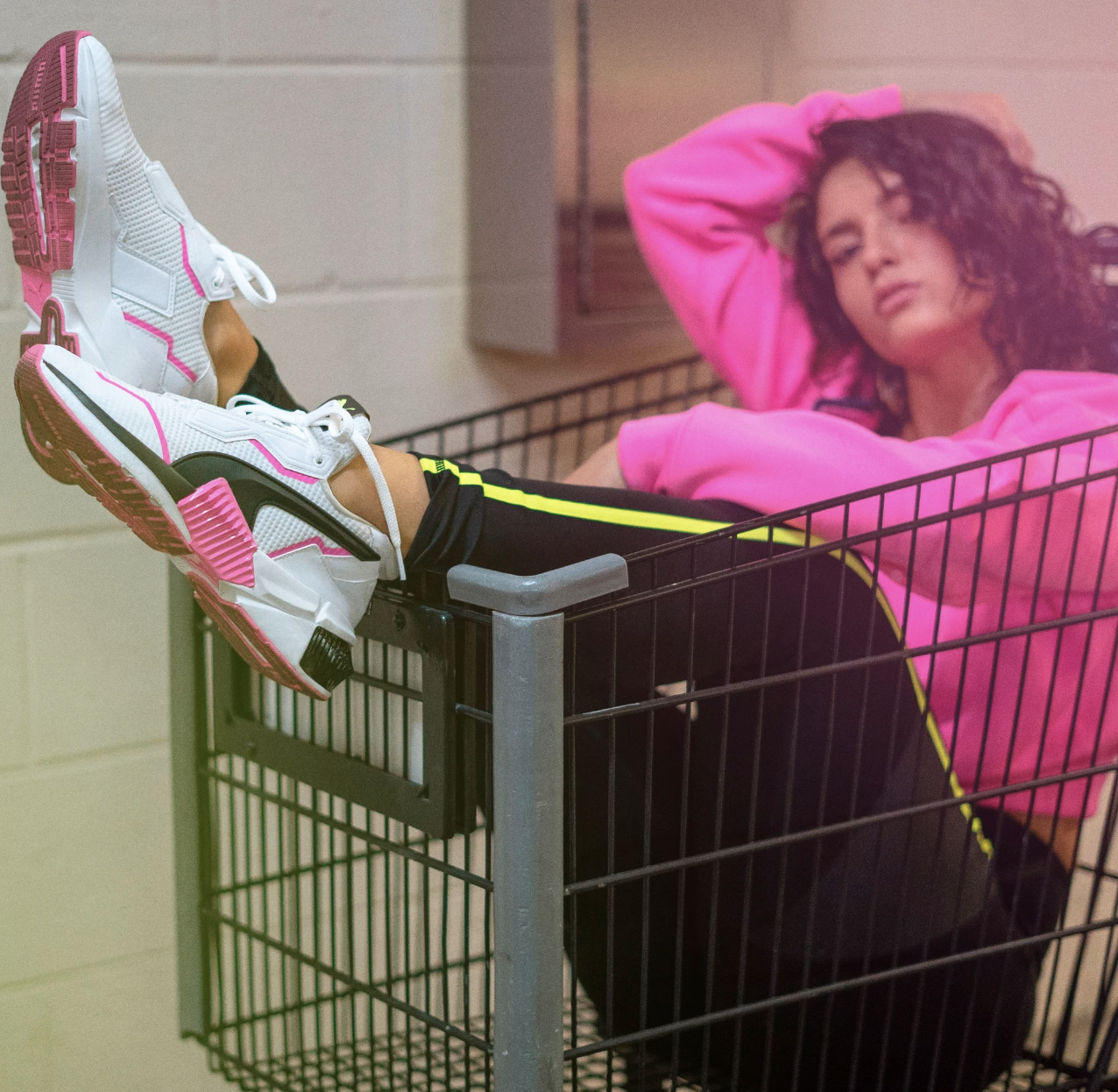 a model sitting in a shopping cart with their feet kicked up to show white, black, and hjot pink sneakers