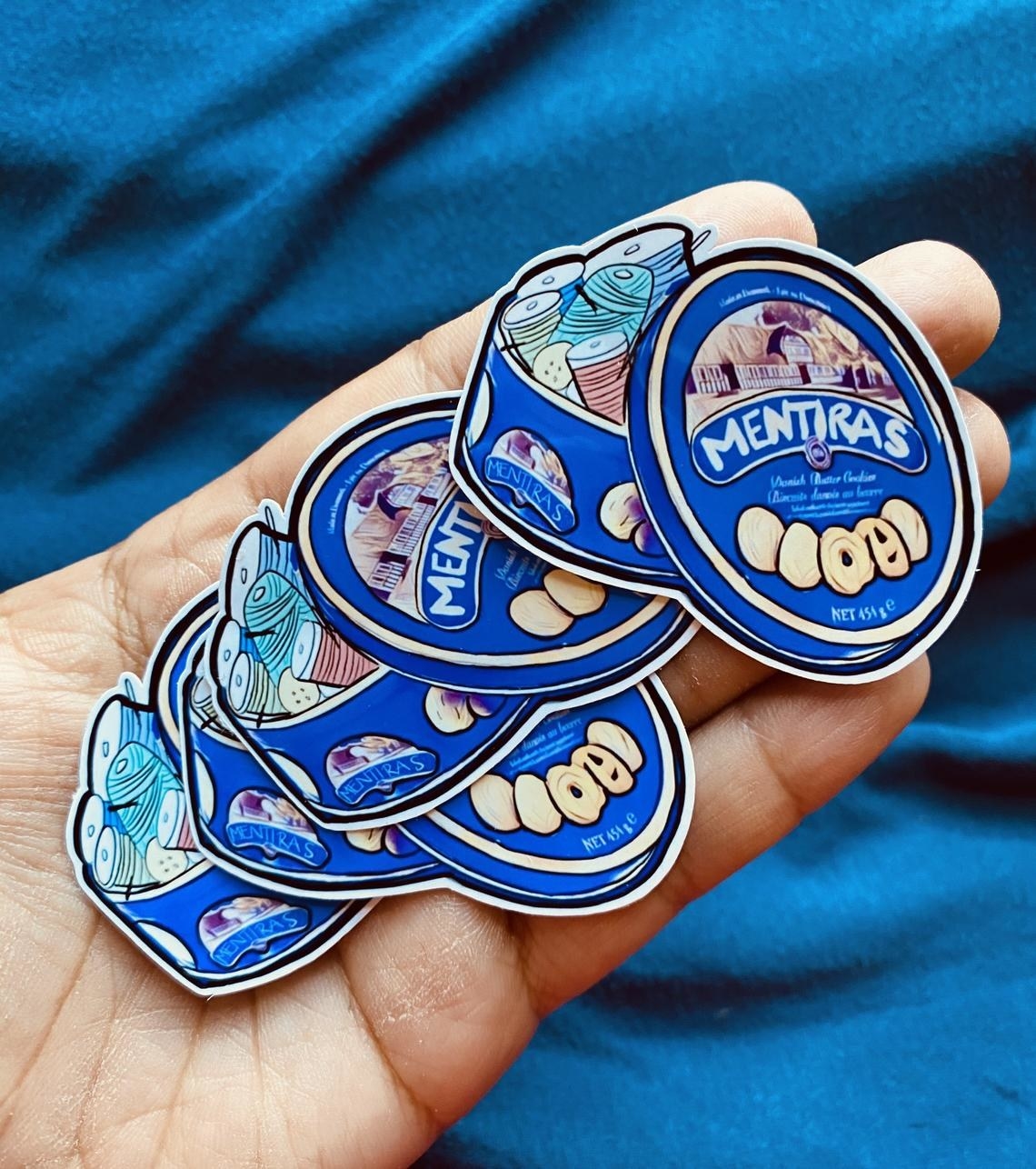 A hand holding a few of the Mentiras blue cookie tin sticker.