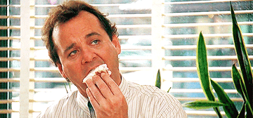 Bill Murray eats a powdered donut while wearing a white striped button-down shirt. He sits in an office and there&#x27;s a snake plant behind him.