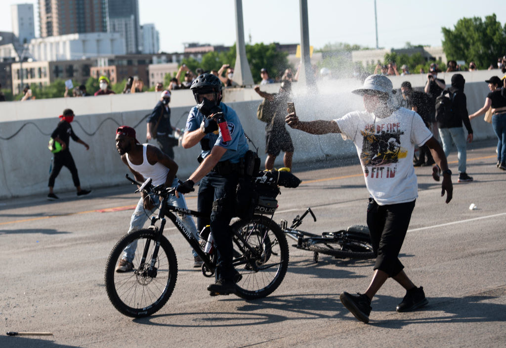 A police officer on a bicycle spraying a protester as he walks down a street