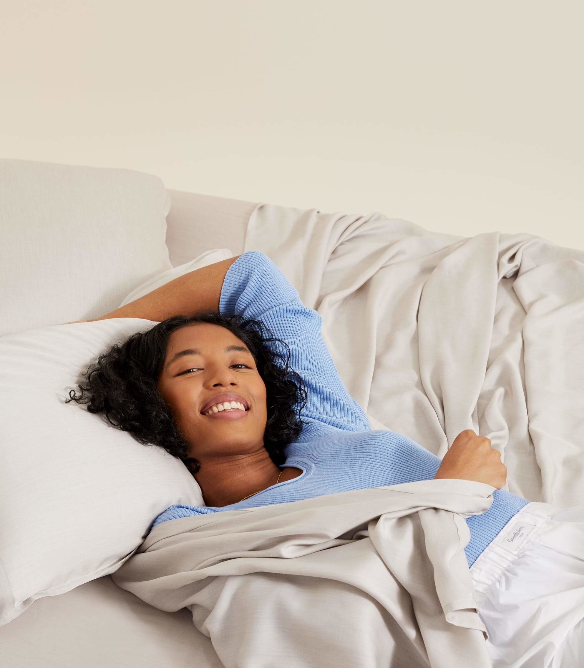 a model smiling and cozy laying on top of light gray sheets