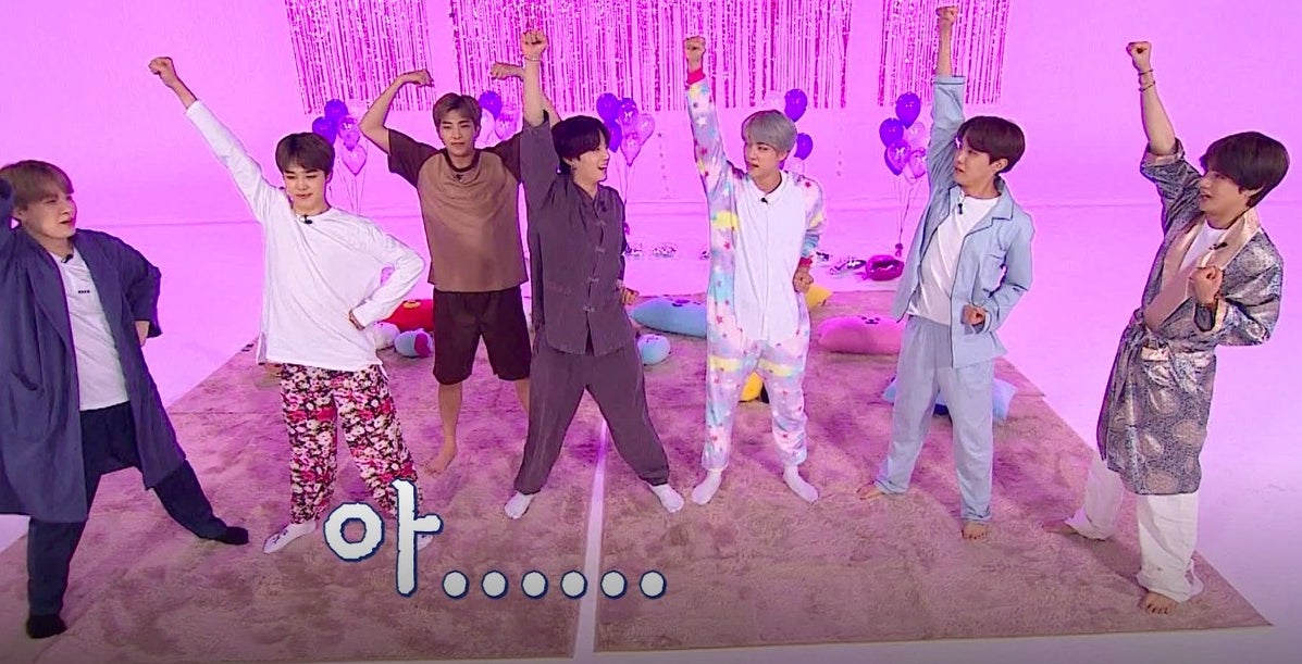 BTS wear pyjamas and have their arms raised on a set decorated with purple streamers and balloons