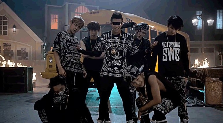 A still from the &quot;No More Dream&quot; music video; BTS pose in front of a school buss with flames surrounding it; they were black and white patterned outfits and lots of jewellery