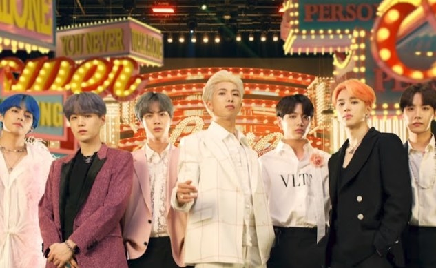 BTS stand in a row wearing formalwear with different coloured hair; there are lit-up signs in the background