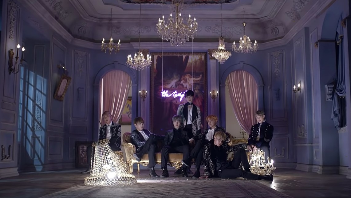 BTS sit in formalwear in a purple elaborately styled room with multiple chandeliers
