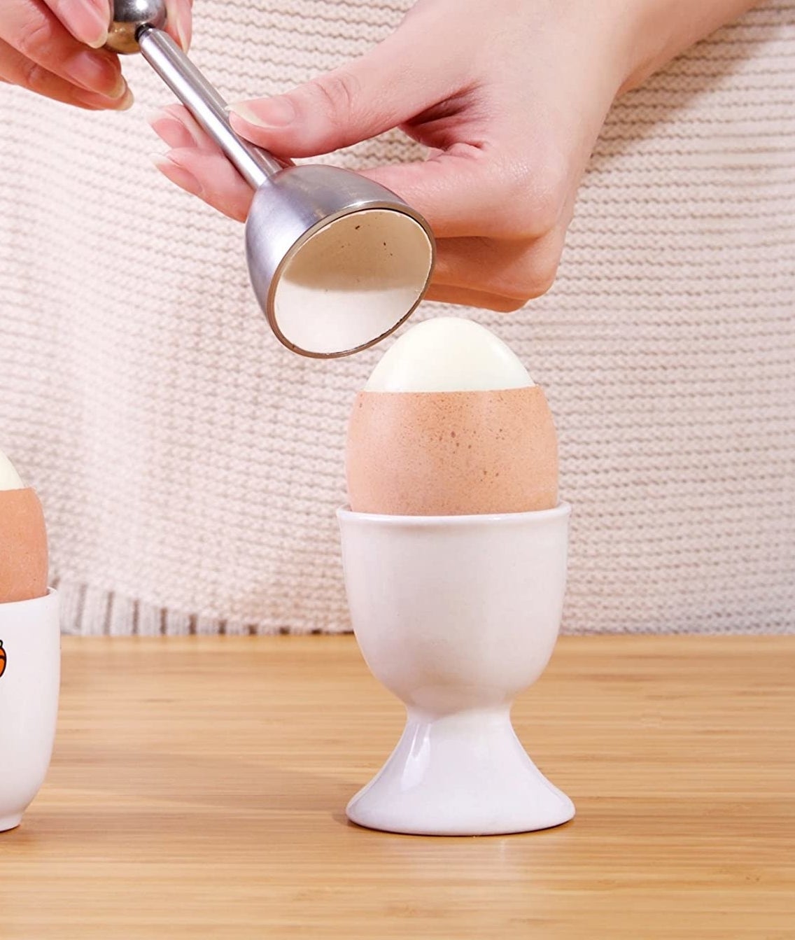 An egg is in a cup on a table with the top of the shell cleanly removed by an egg cracker being held in two hands