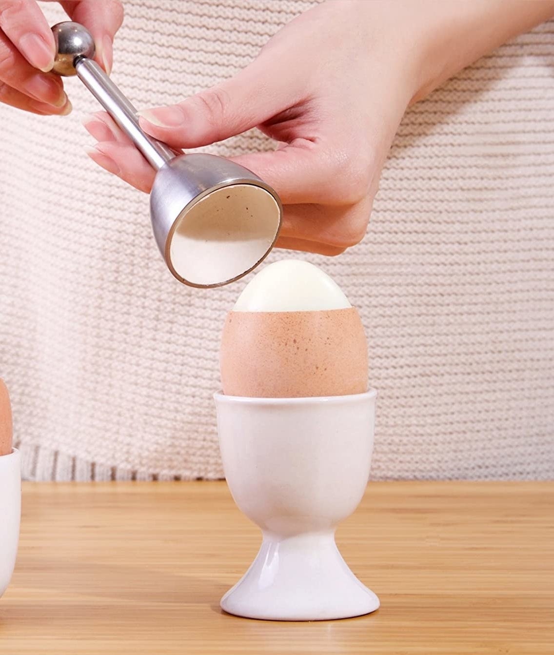 An egg is in a cup on a table with the top of the shell cleanly removed by an egg cracker being held in two hands