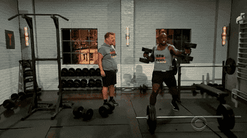 A GIF of an astonished James Corden in the gym with Terry Crews who is working out and dancing with heavy dumbells