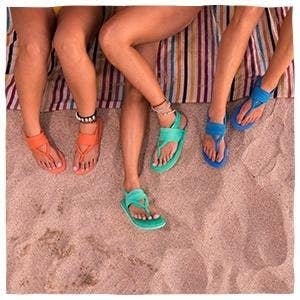 Three models wearing different colored Sanuk sling sandals on the beach.
